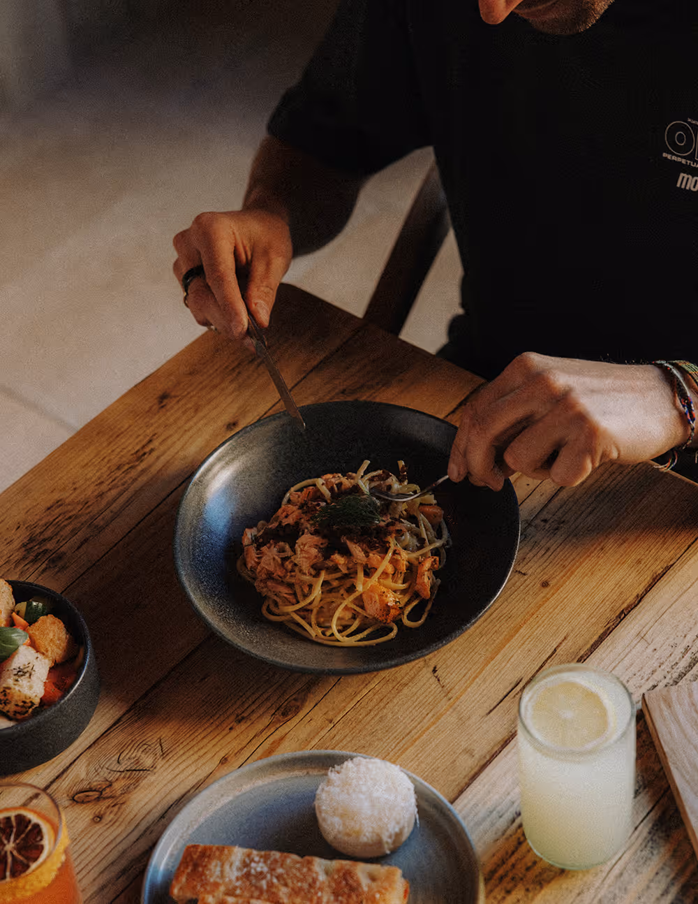 Person eating spaghetti with fork and knife at a wooden table with salad, bread, and drinks.