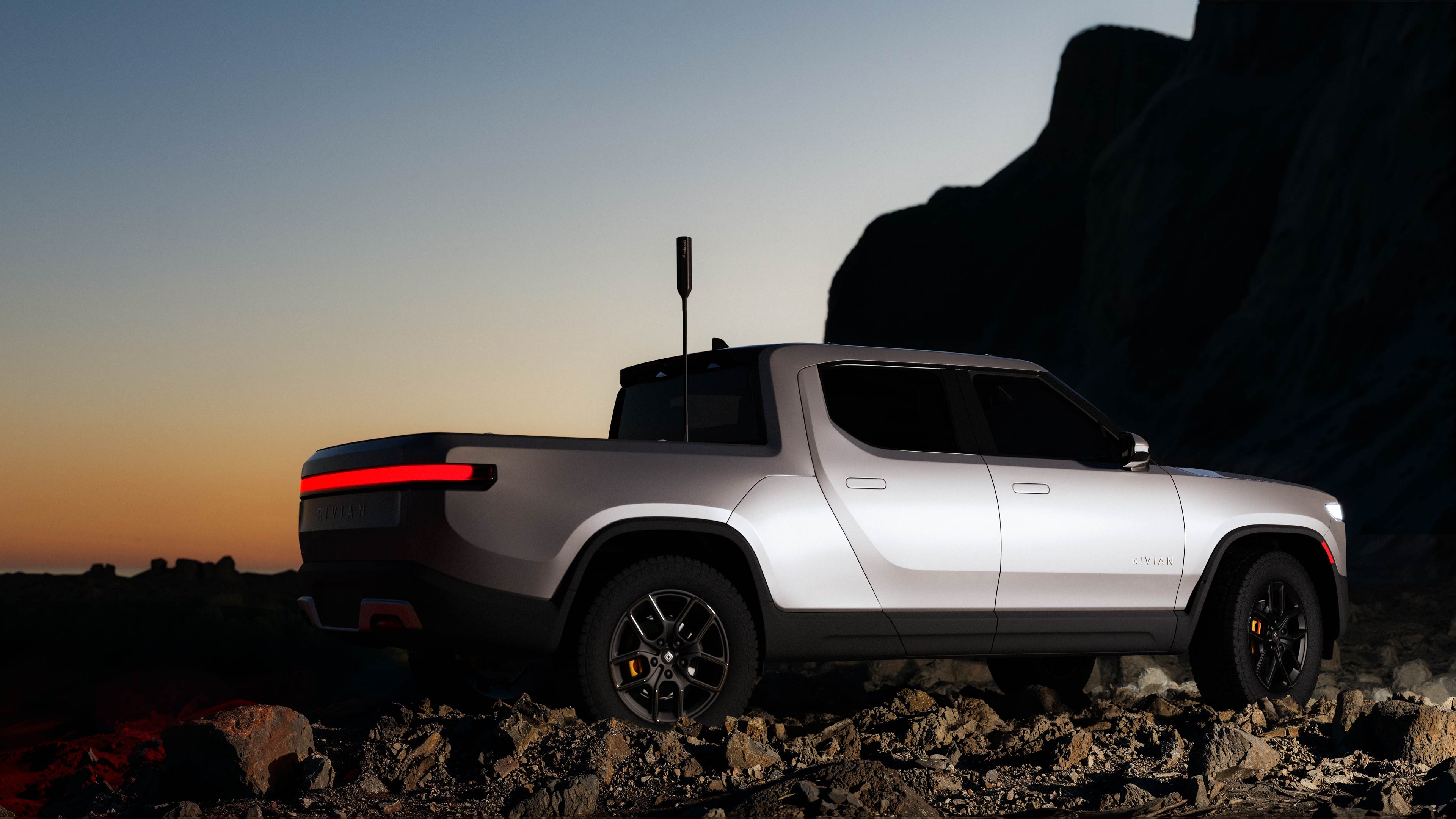 We Boost antenna on a Silver Rivian electric pickup truck parked on rocky terrain at sunset.