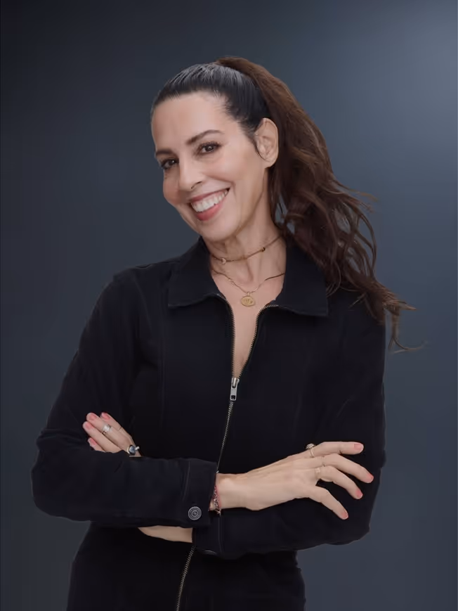Smiling woman with long dark hair in a ponytail, wearing a black zip-up jacket and layered necklaces, standing with arms crossed.