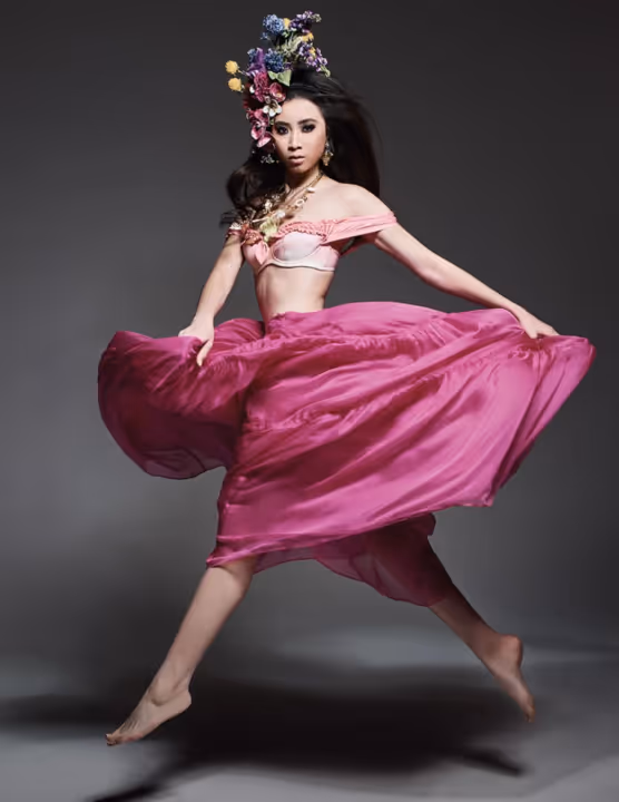 Woman in a flowing pink skirt and floral headpiece gracefully jumping against a gray background.