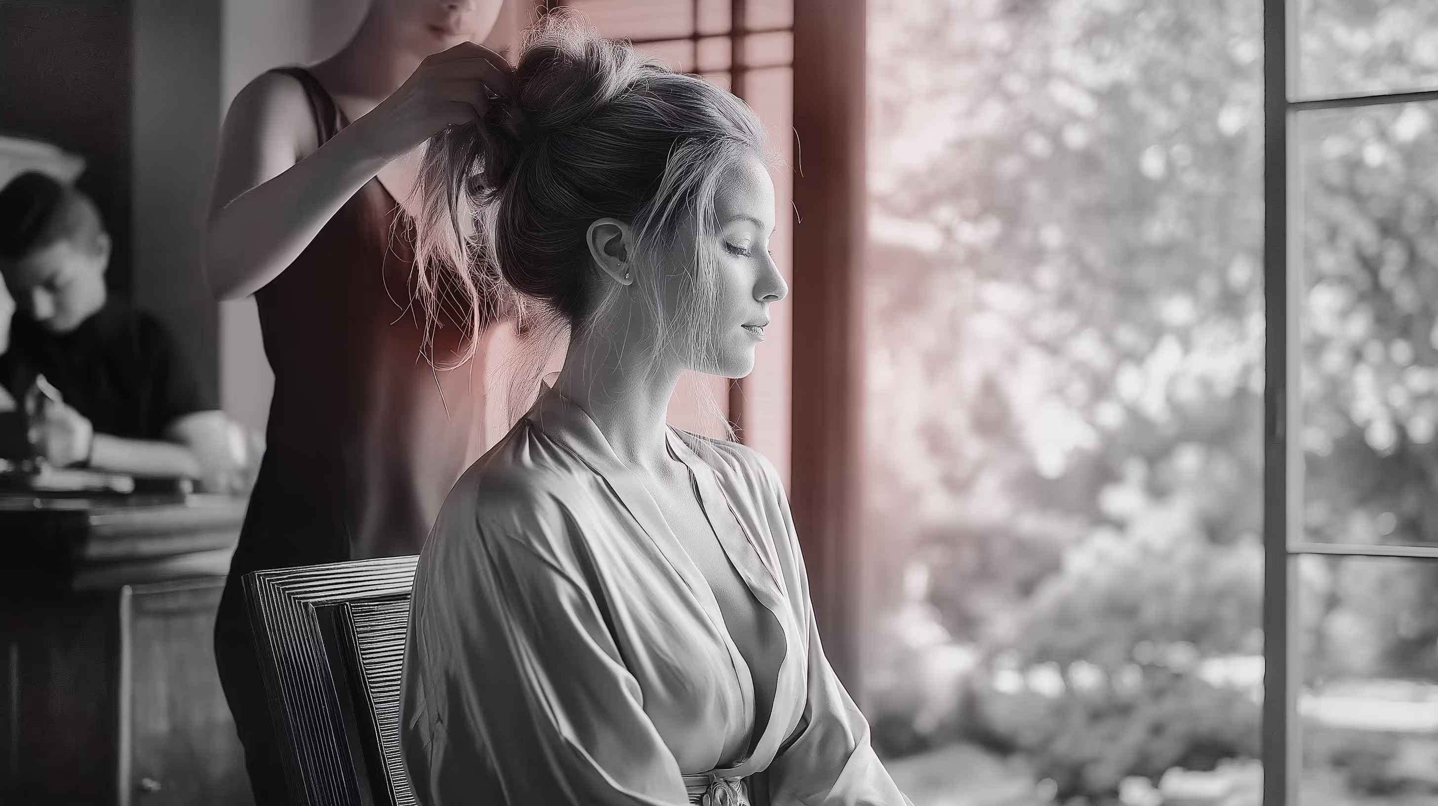 Woman sitting with eyes closed while another person styles her hair in a softly lit room near a window.