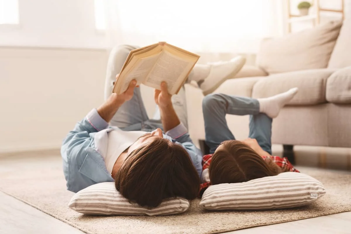 Apa és lánya a nappaliban fekszenek a földön, miközben könyvet olvasnak együtt.