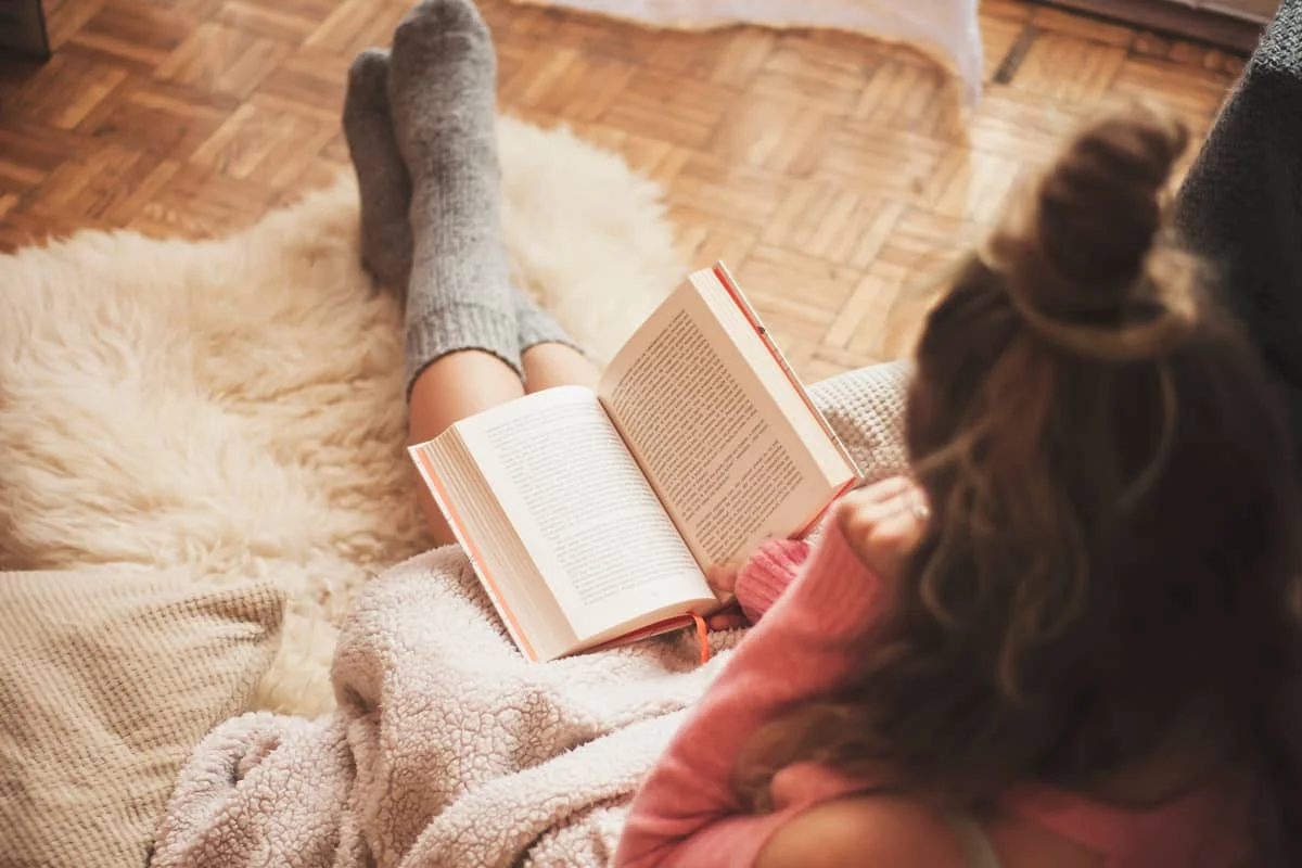O femeie, înfășurată într-o pătură caldă, stă pe covor în șosete groase și citește o carte într-un mediu calm și familial.