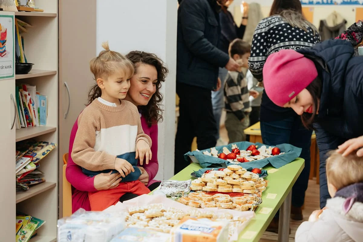 Anyuka kisfiával az óvoda adventi jótékonysági vásárán, ahol házi sütemények és karácsonyi finomságok sorakoznak az asztalon.