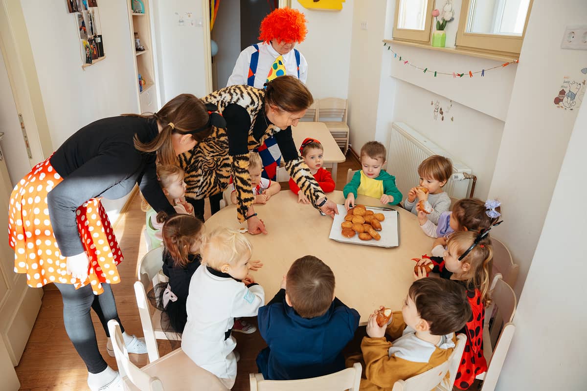 Bölcsődés gyermekek farsangi jelmezben farsangi fánkot esznek.