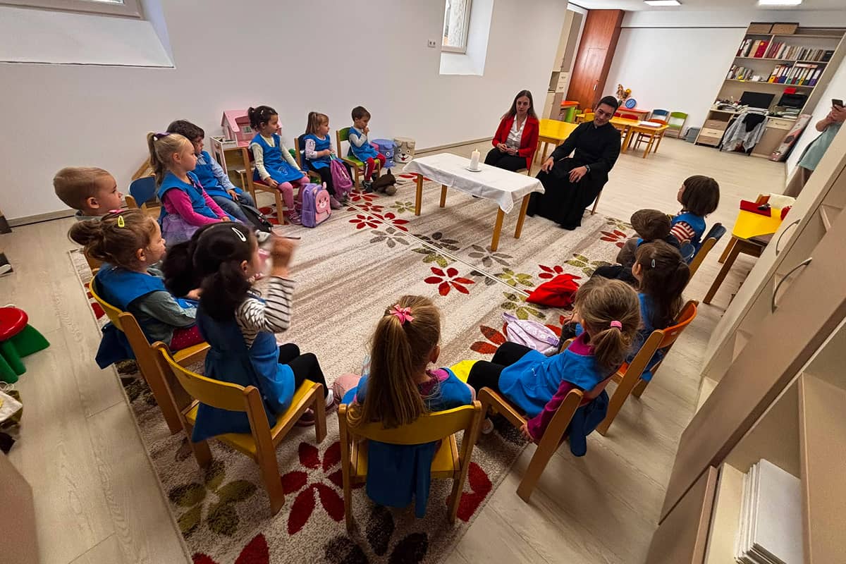 Óvodás gyermekek táskáikkal maguk mellett hallgatják Sebestyén Izabella hitoktatót és Bege Lóránd kispapot a táskaszentelés alkalmával.