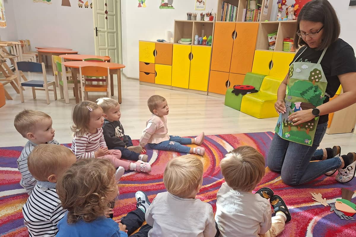 Bölcsődés gyermekek figyelmesen hallgatják a népmesét, miközben az óvónő mesekötény segítségével jeleníti meg a történet szereplőit.