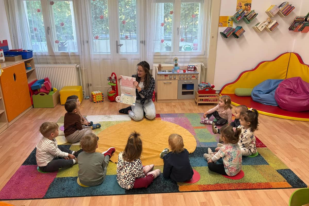 Bölcsődés gyermekek körben ülve figyelmesen hallgatják a mesét, miközben az óvónő képes mesélőeszközzel szemlélteti a történetet.