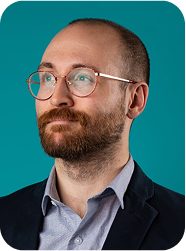 Portrait d’un homme barbu avec lunettes, vêtu d’une chemise bleue et veste foncée, sur fond bleu turquoise.