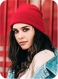 Portrait d’une femme portant un bonnet rouge et une veste en jean, posant devant un fond rouge vif.