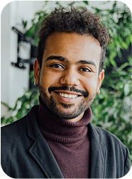 Portrait d’un homme souriant portant un col roulé bordeaux et une veste sombre, posé devant un fond végétal.