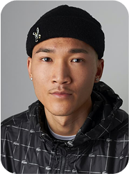 Portrait d’un homme portant un bonnet noir et une veste à carreaux sombres, posant sur fond gris neutre.