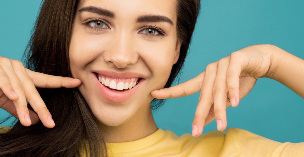 Brunette vrouw met haar handen en vingers wijzend naar haar glimlach voor een blauwe achtergrond