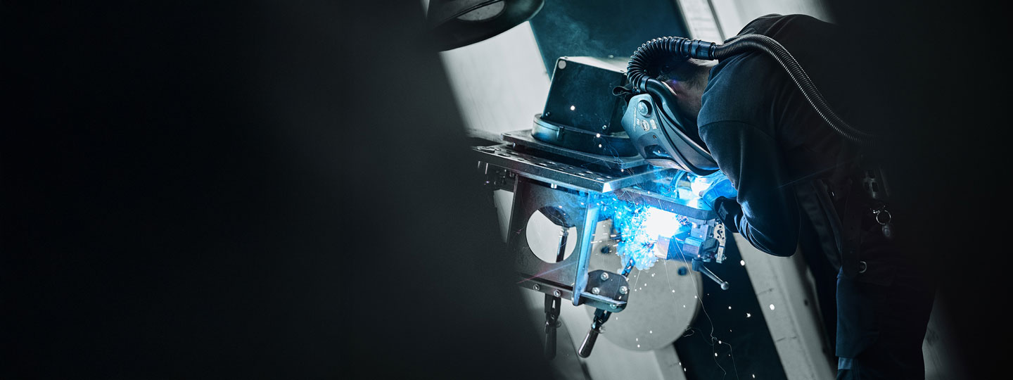 Welder wearing protective gear welding a metal structure with bright blue sparks.