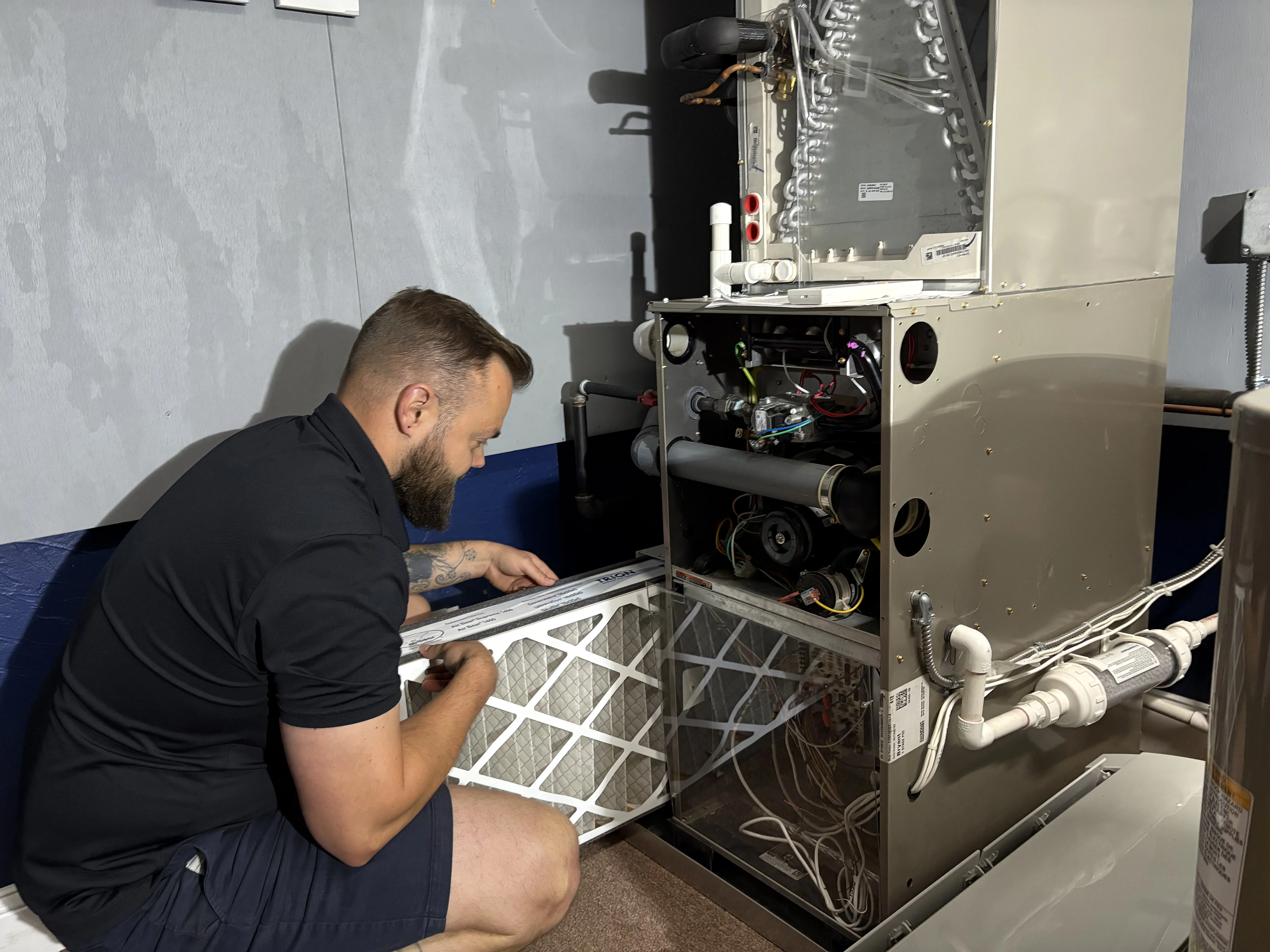 Man in black shirt and shorts kneeling and installing a furnace air filter into an open HVAC unit.