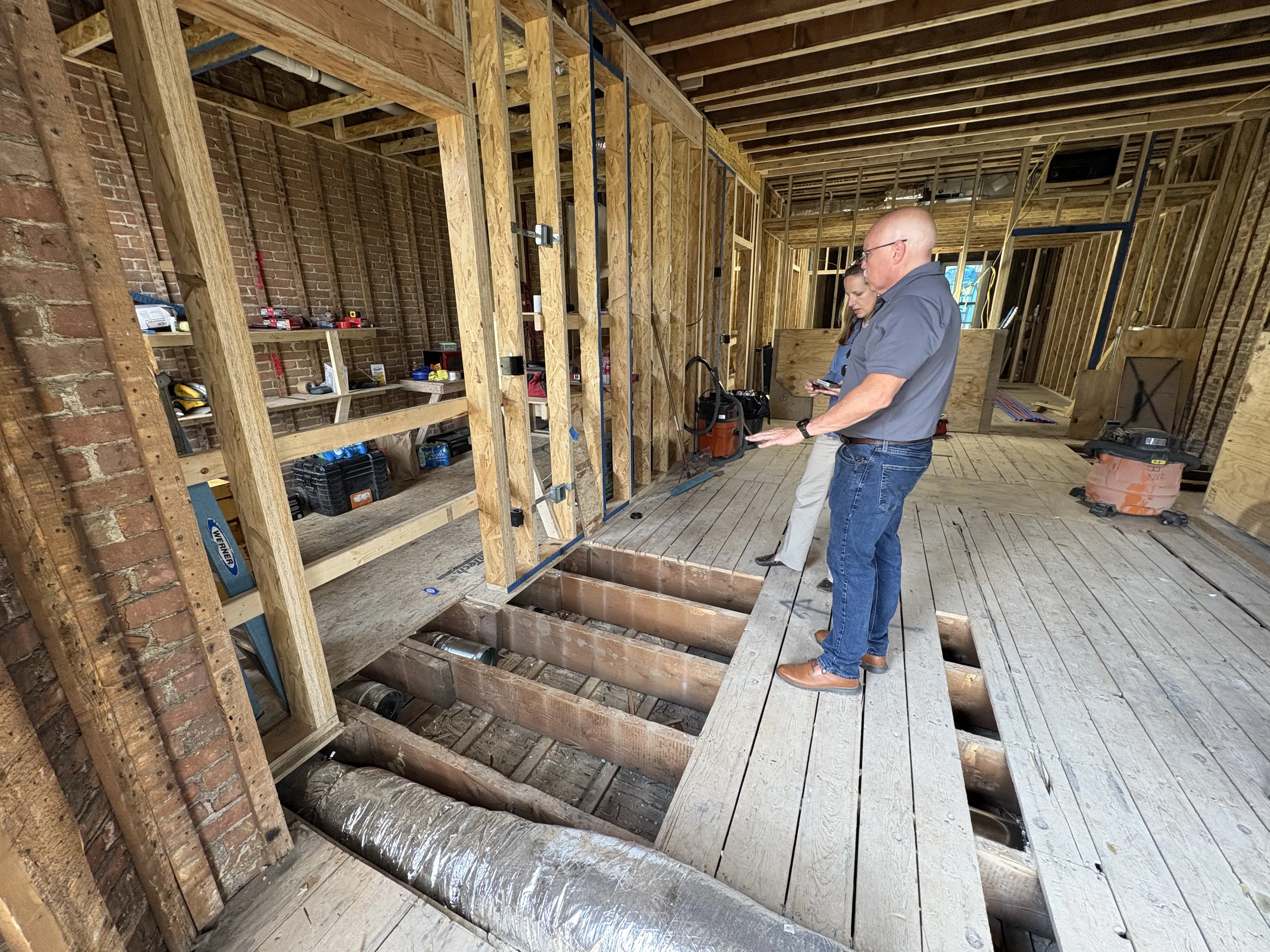 Two people inspecting an indoor construction site with exposed wooden floor joists and framework.