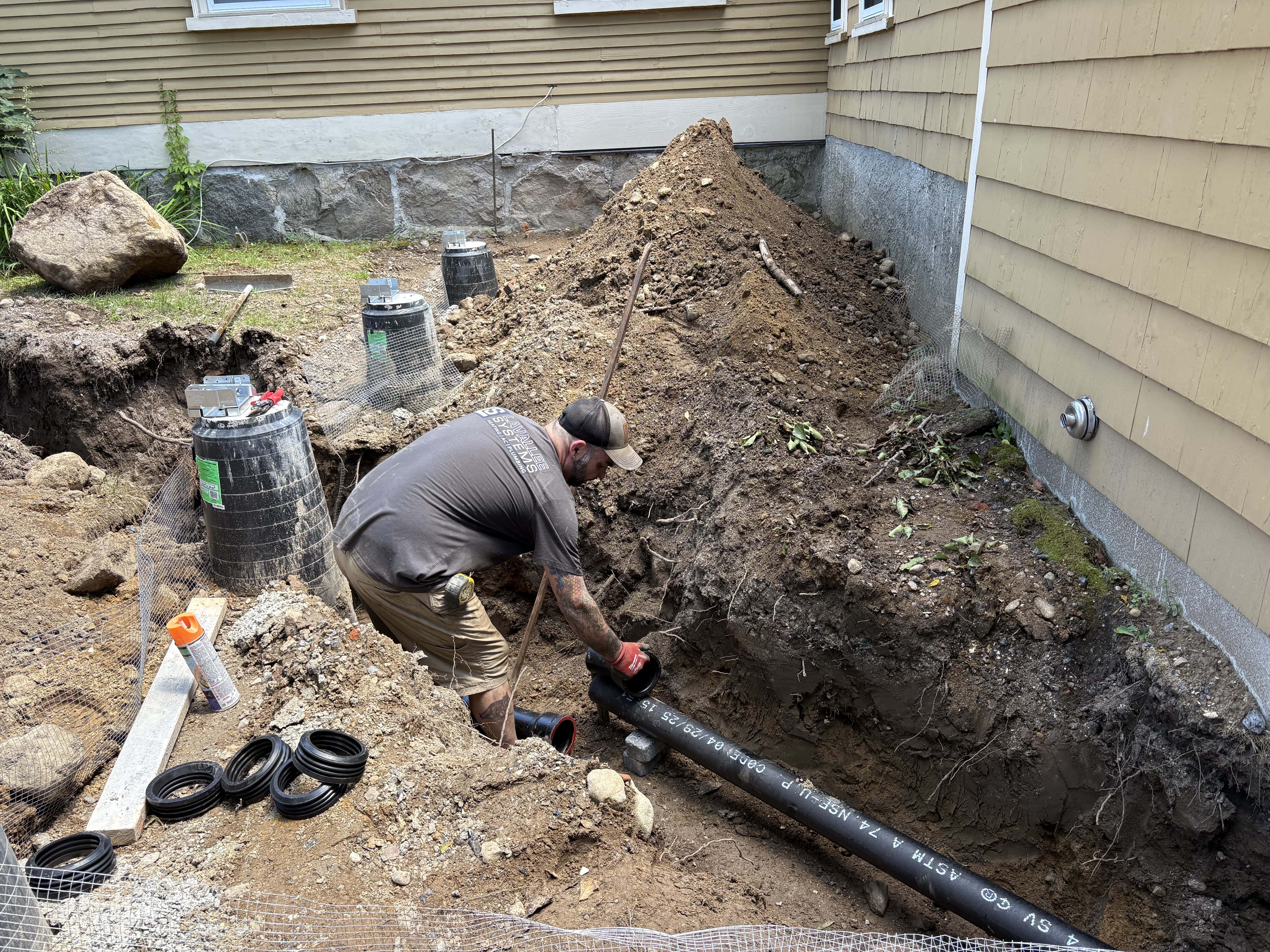 Worker installing black underground drainage pipes in a dug trench outside a house with dirt piles around.
