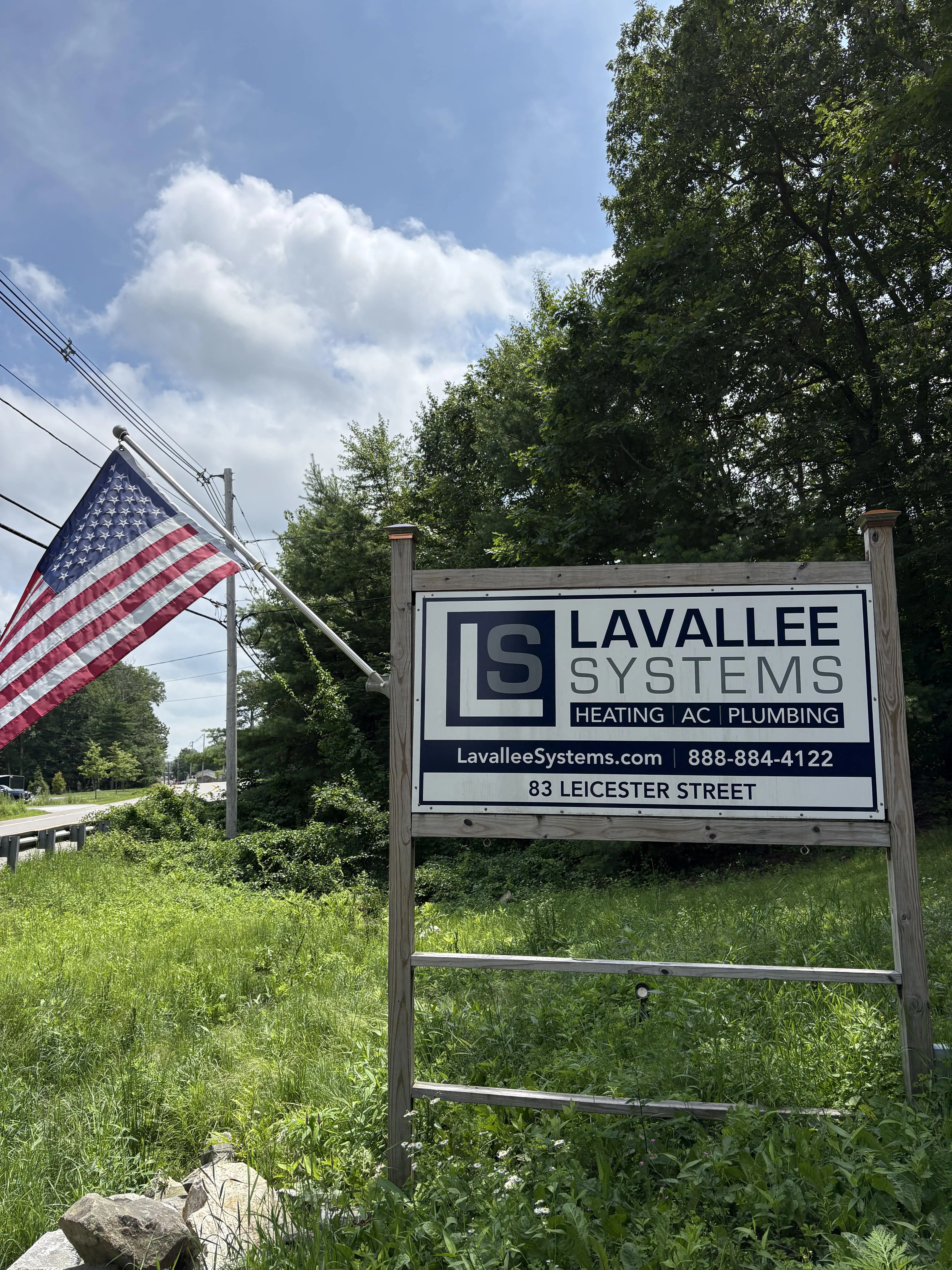 Outdoor sign for Lavallee Systems heating, AC, and plumbing at 83 Leicester Street with an American flag on a pole beside it.