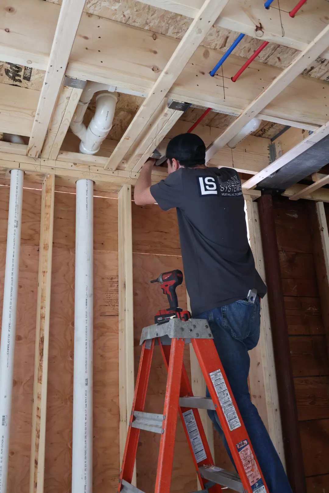 Worker in a black LS Systems shirt standing on an orange ladder installing pipes in a wooden framed ceiling.
