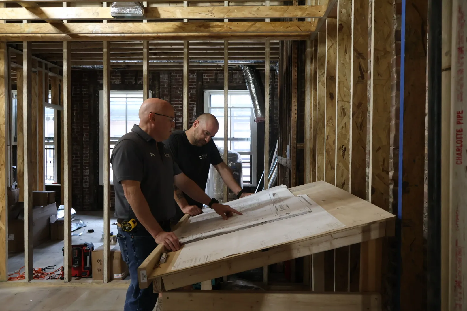 Two men reviewing blueprints on a wooden drafting table inside a partially constructed building with exposed framing and pipes.