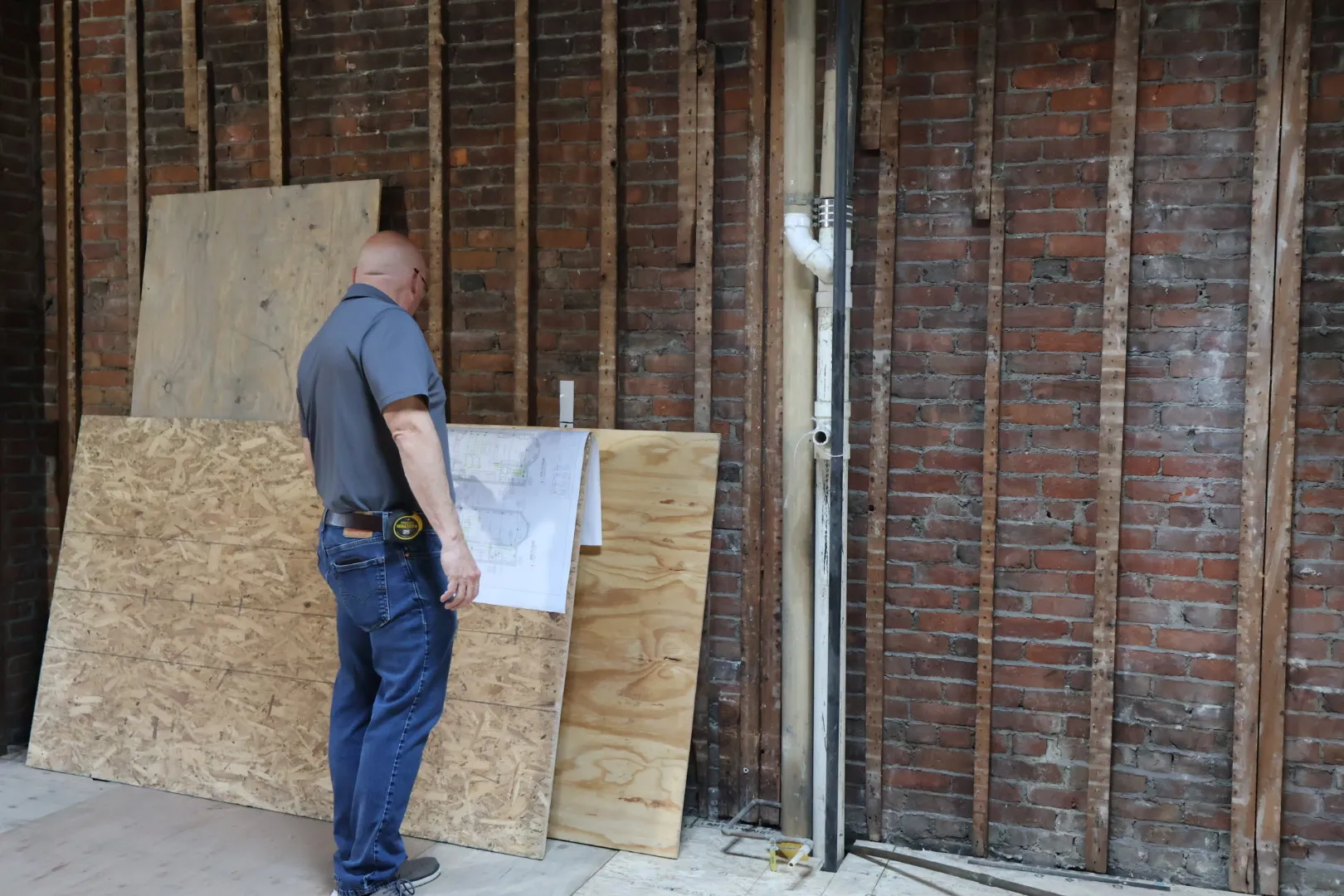 Man in jeans and gray shirt examining blueprints in a room under renovation with exposed brick walls and wooden panels.