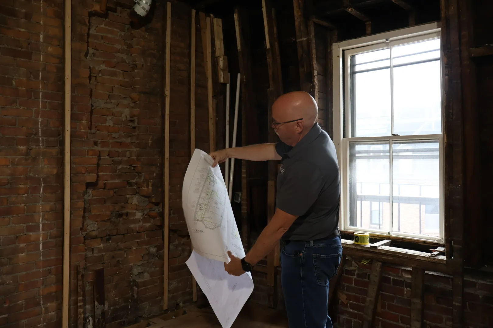 Bald man in glasses and gray shirt examining architectural plans inside a partially renovated brick room with an exposed window.