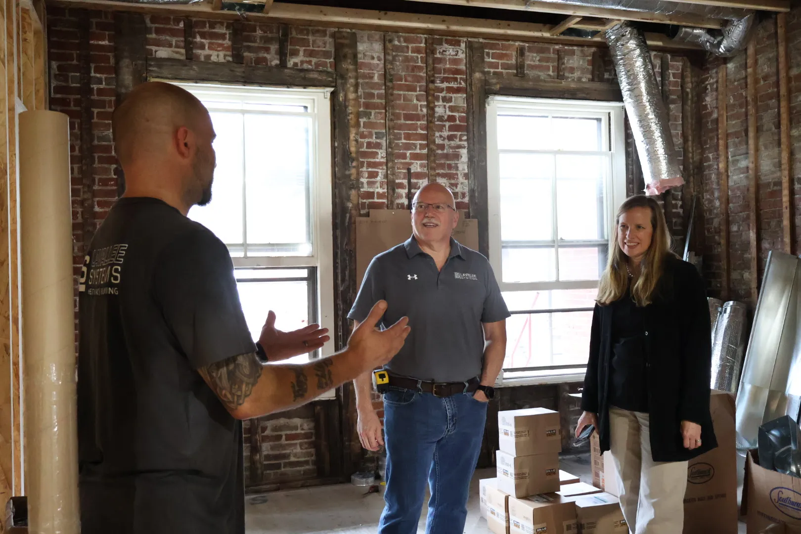 Three people having a discussion inside a partially renovated room with exposed brick walls and stacked cardboard boxes.