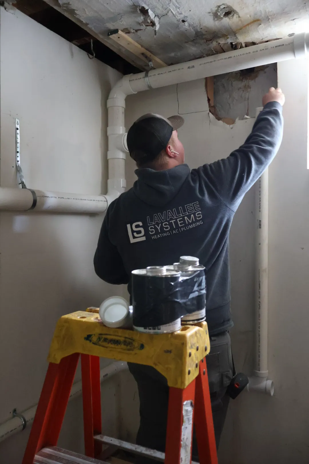 Person wearing a Lavallee Systems hoodie inspecting plumbing pipes in a room near a window.