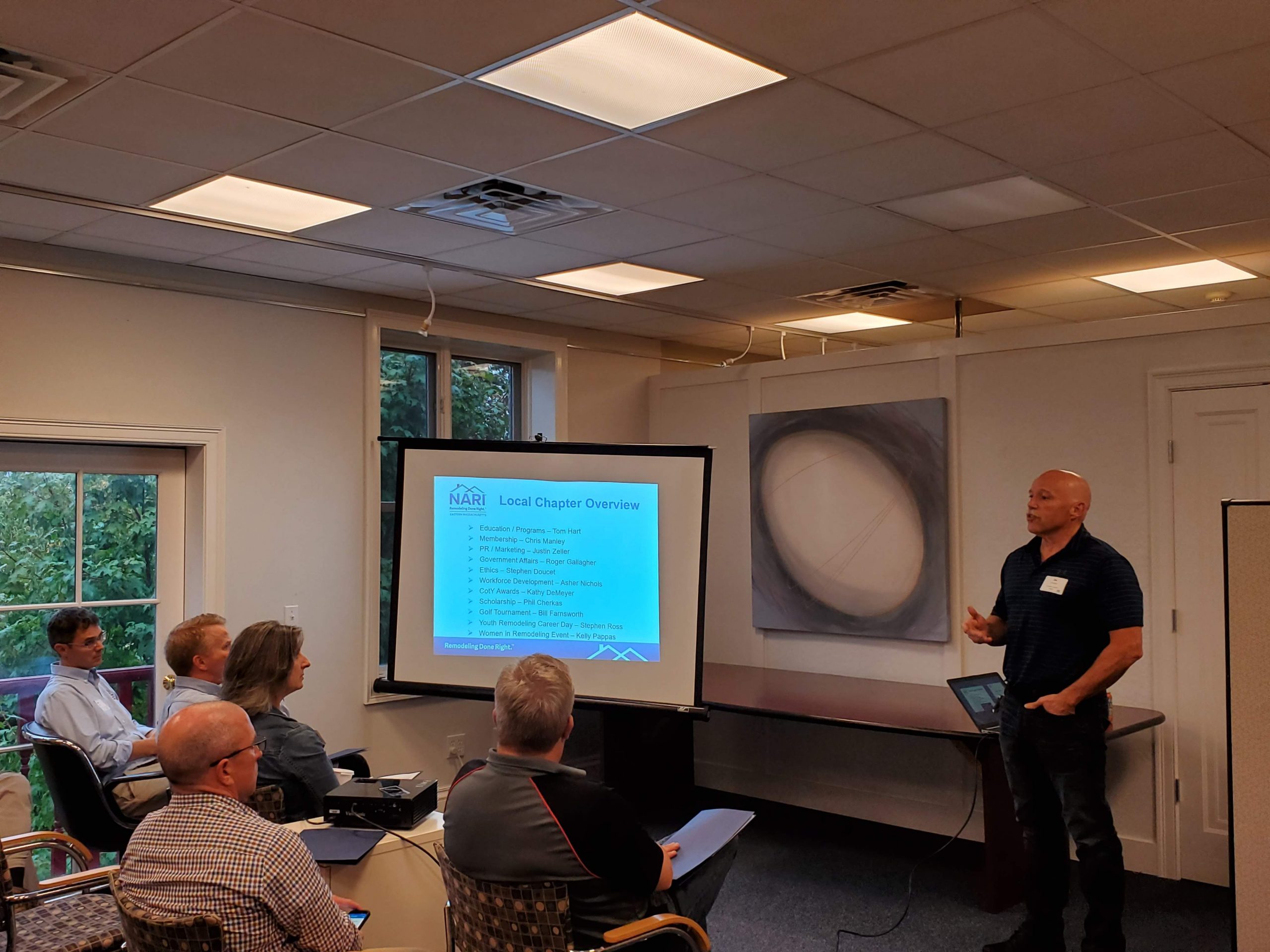 Man giving a presentation to five seated attendees in a conference room with a projector screen showing a local chapter overview.