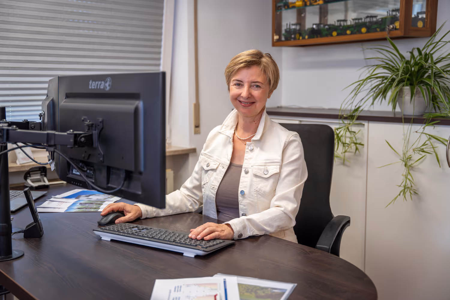 Hildegard Zeltsperger trägt eine weiße Jack und sitzt in einem Büro von Zeltsperger. Brauner Bürotisch mit Desktops, Unterlagen und einer Tastatur.