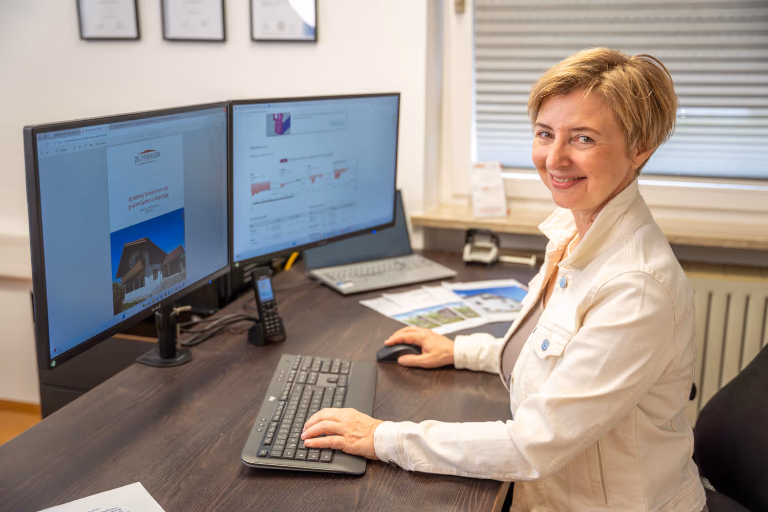 Hildegard Zeltsperger trägt eine weiße Jack und sitzt in einem Büro von Zeltsperger. Brauner Bürotisch mit Desktops, Unterlagen und einer Tastatur.