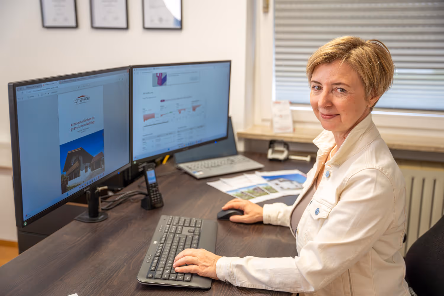 Hildegard Zeltsperger trägt eine weiße Jack und sitzt in einem Büro von Zeltsperger. Brauner Bürotisch mit Desktops, Unterlagen und einer Tastatur.