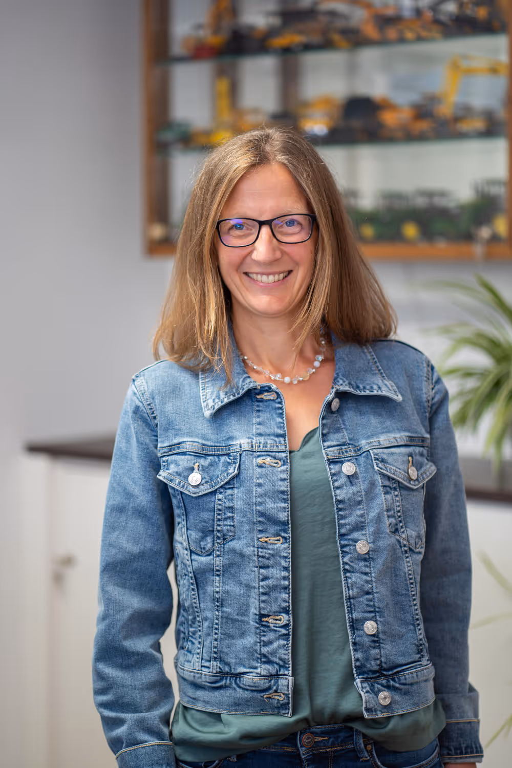 Andrea Zierer trägt eine Jeansjack und steht in einem Büro mit einer Pflanze.