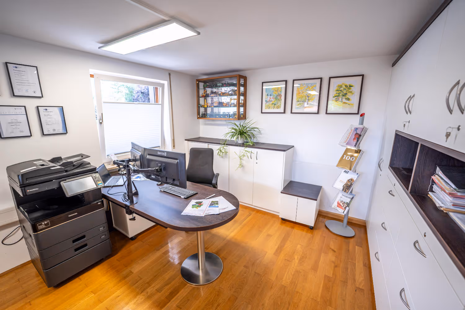 Helles Büro von Zeltsperger Immobilien und Wirtschaftsberatung mit Auszeichnungen an der Wand, braunem Bürotisch und Desktops und Tür mit Regalen.
