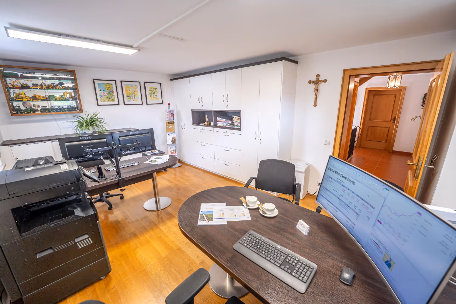 Helles Büro von Zeltsperger Immobilien und Wirtschaftsberatung mit Auszeichnungen an der Wand, braunem Bürotisch und Desktops und Tür mit Regalen.