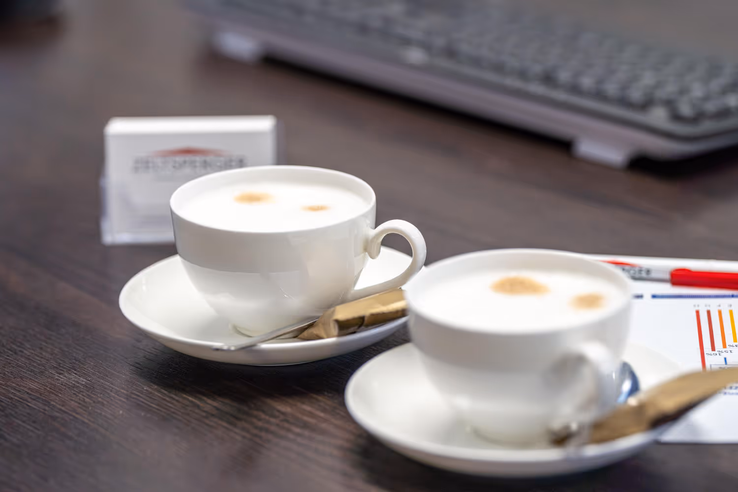 Zwei Kaffeetassen mit Zucker stehen auf einem braunen Tisch mit Visitenkarten und einer Tastatur.