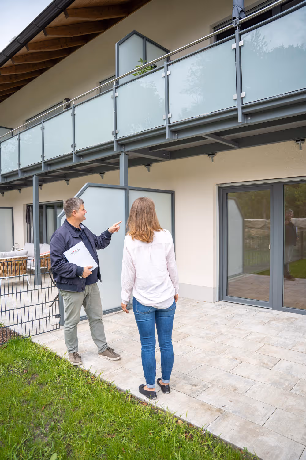 Hubert Zeltsperger, ein Mann mittleren alters steht vor einem Haus im Garten und erklärt einer blonden Frau in Jeans etwas über das Haus.