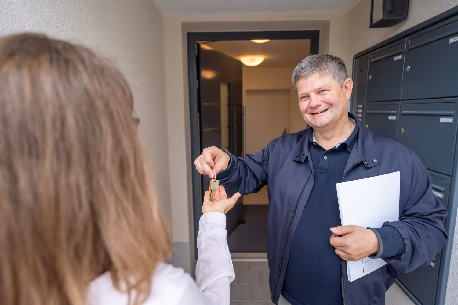 Hubert Zeltsperger überreicht einen Schlüssel an eine Frau. Im Hintergrund eine offene Tür und Briefkästen.