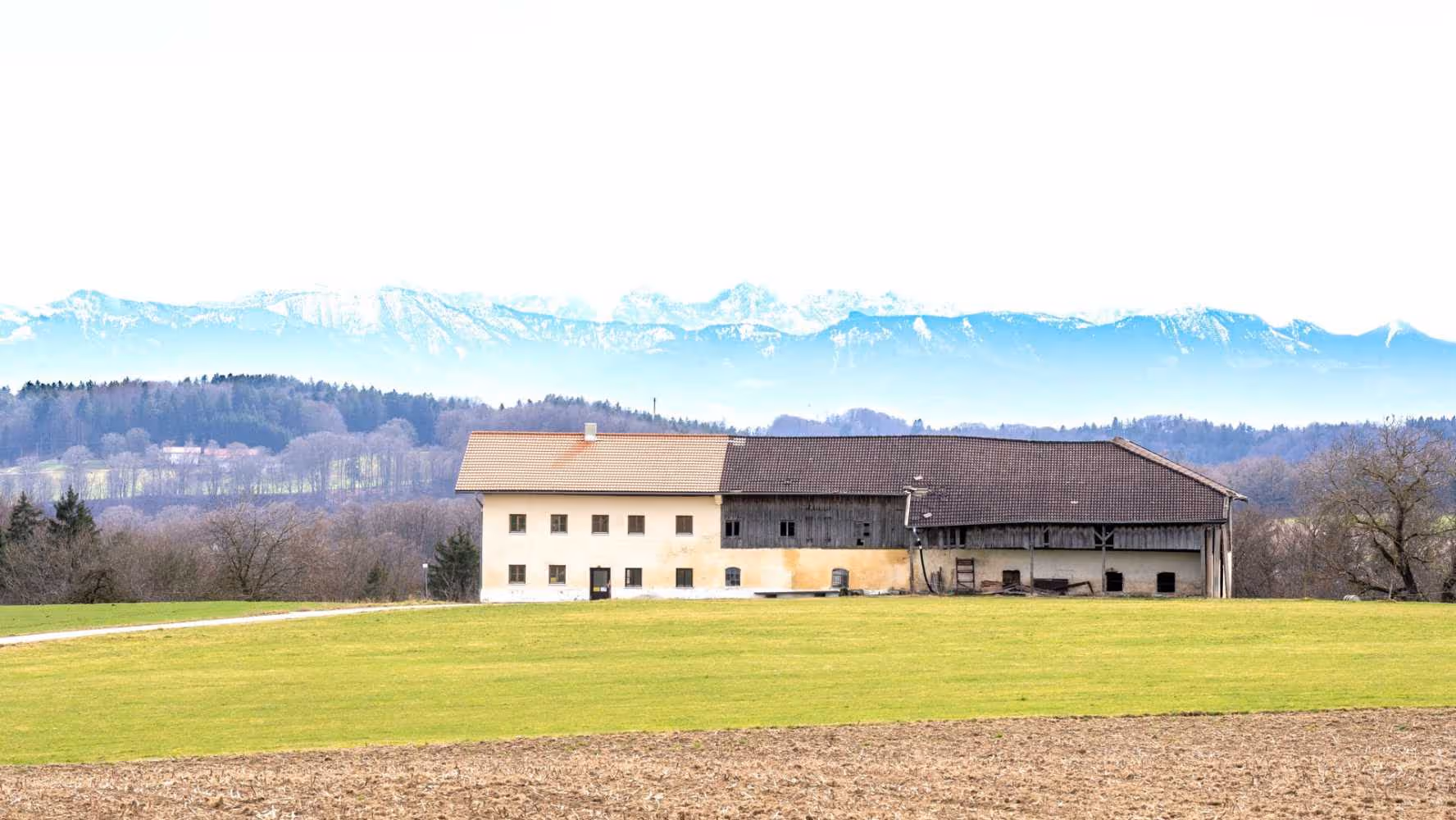Traditioneller Bauernhof auf einer Wiese mit Alpenpanorama im Hintergrund.