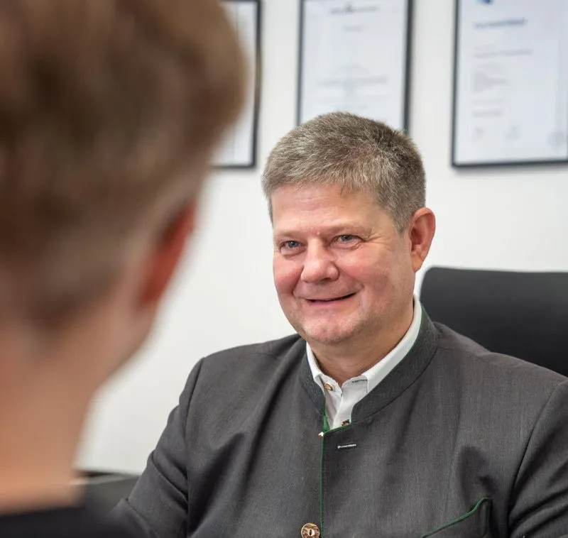 Hubert Zeltsperger unterhält sich mit einer Person. Im Hintergrund an der Wand hängen Zertifikate.
