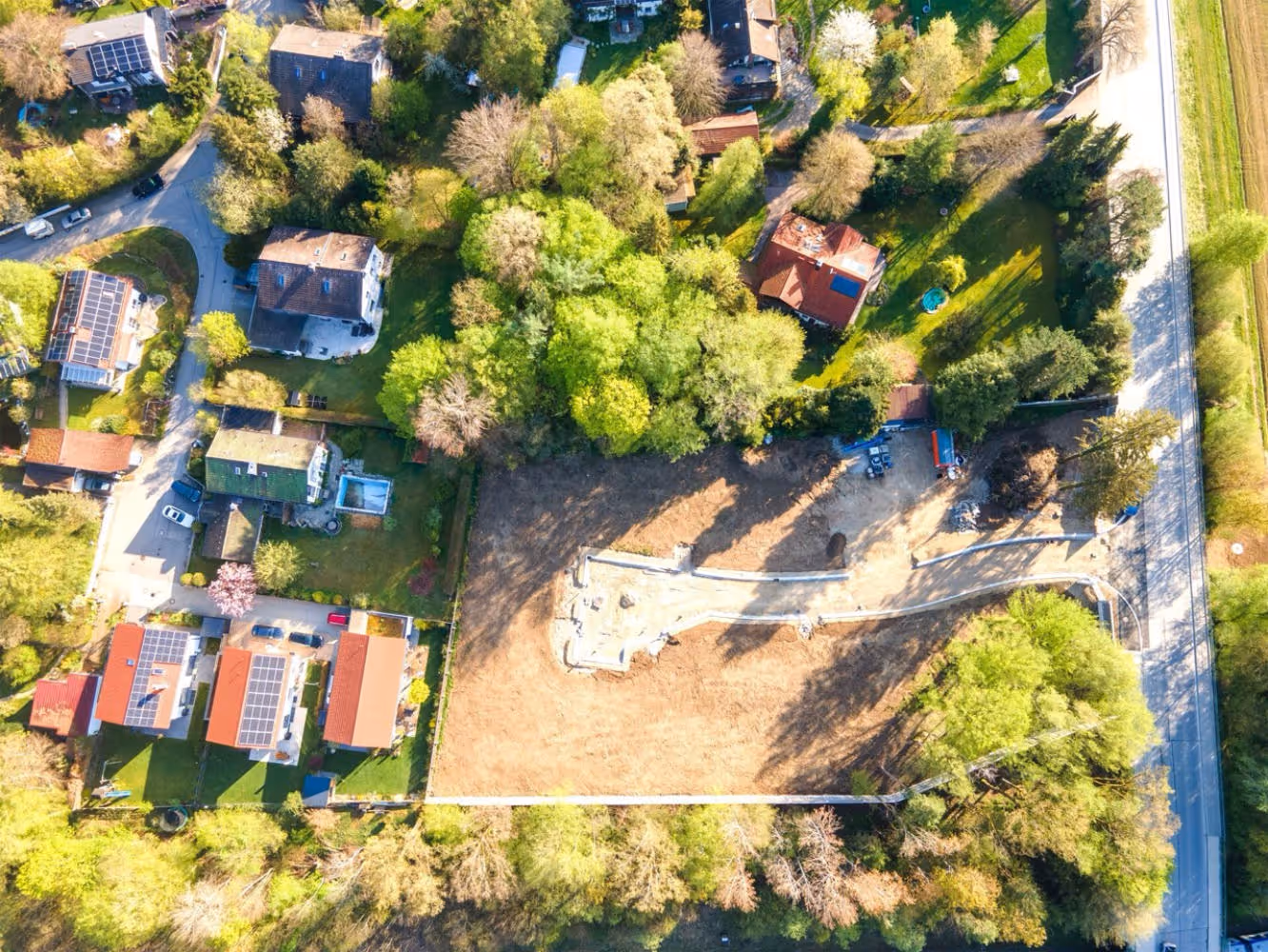 Luftaufnahme eines Baugrundstücks mit Fundamentarbeiten, umgeben von Bäumen, Straßen und einem Bahngleis.