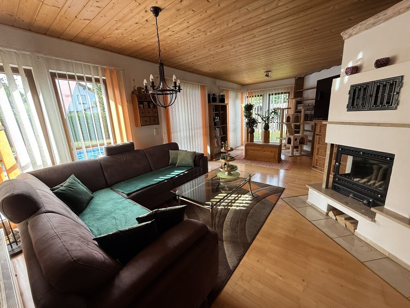 Geräumiges Wohnzimmer mit Parkettboden, großer Ecksofa in Grau, Kaminofen, bodentiefen Fenstern mit Blick ins Grüne und offener Essbereich – helles, gemütliches Einfamilienhaus-Interieur.