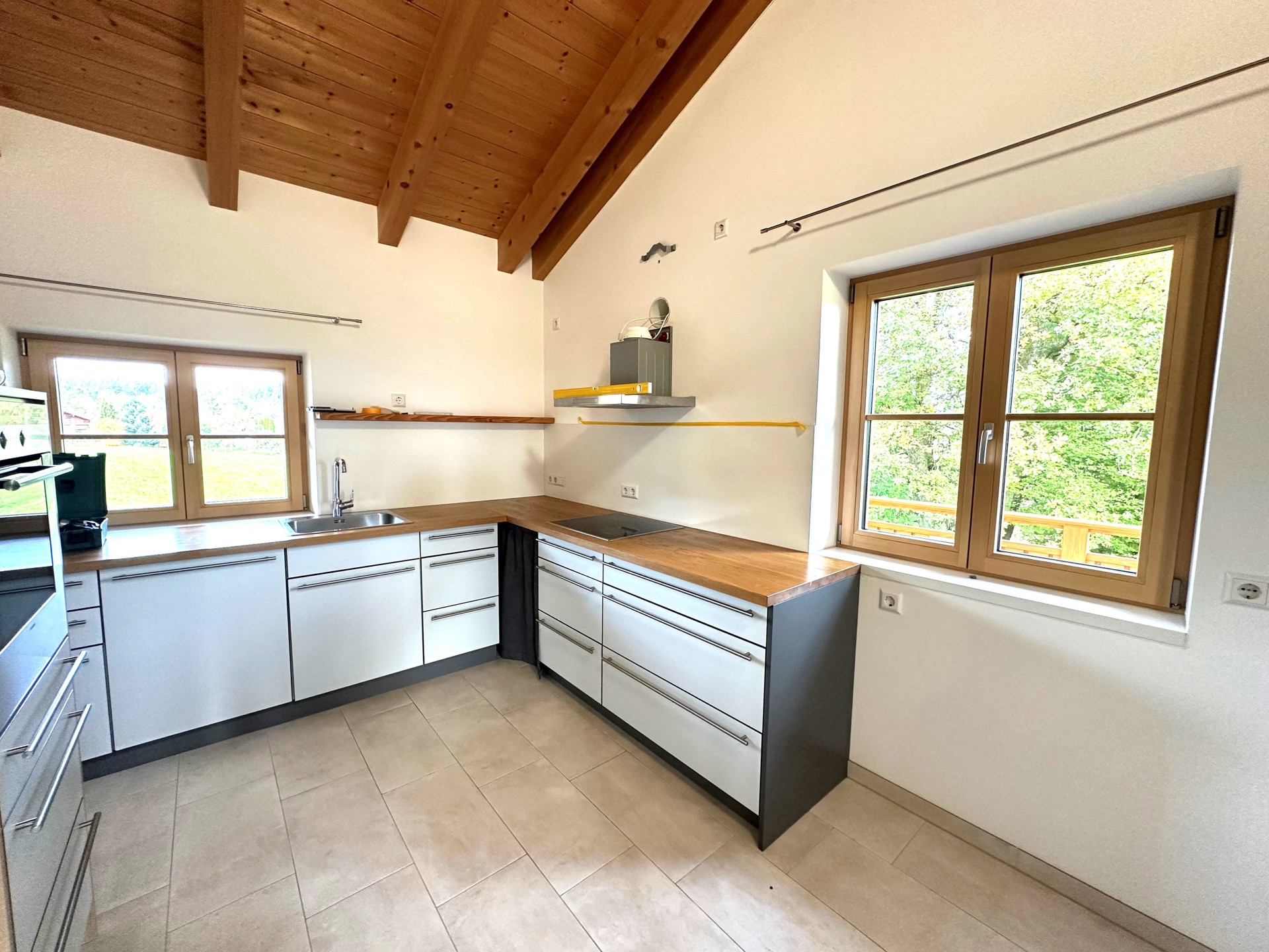 Helle moderne Küche mit weißem Mobiliar, Holzarbeitsplatten, großen Fenstern und sichtbarem Holzgebälk unter der Dachschräge.