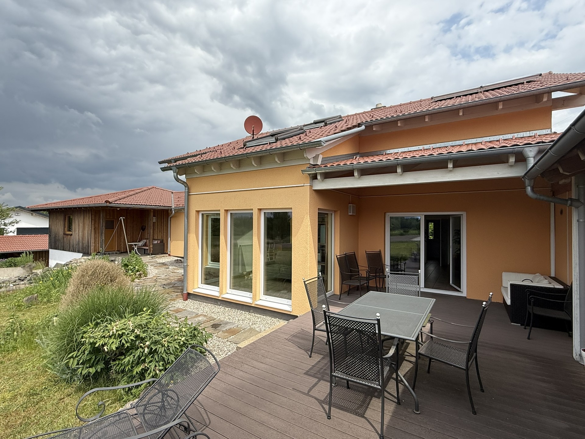 Sonnige Terrasse mit Esstisch und Stühlen vor einem modernisierten orangefarbenen Haus mit großen Fensterfronten und Markise, umgeben von gepflegtem Garten.