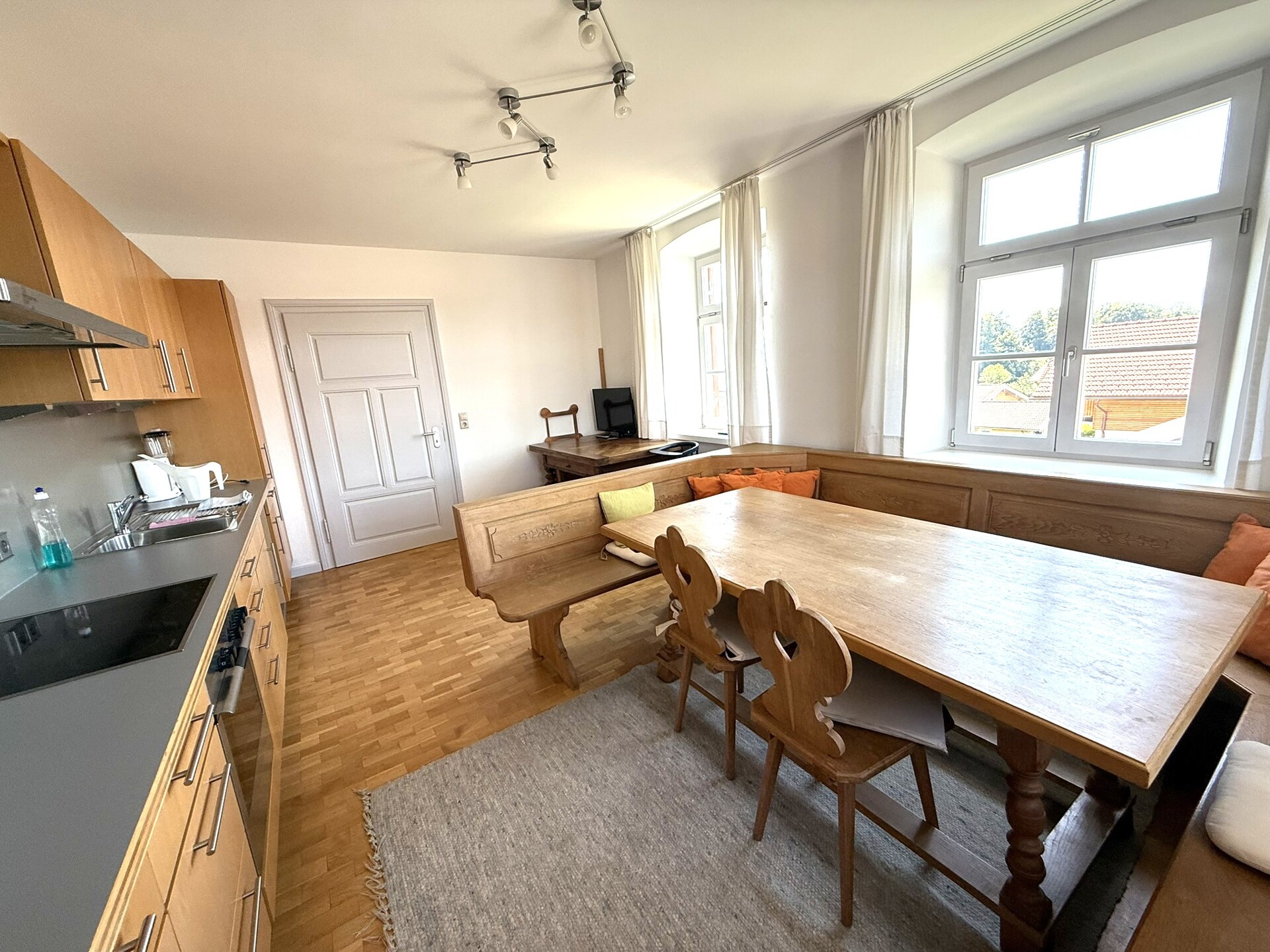 Helle Wohnküche mit großem Holzesstisch, Eckbank und moderner Einbauküche; Tageslicht fällt durch zwei große Fenster in den Raum.