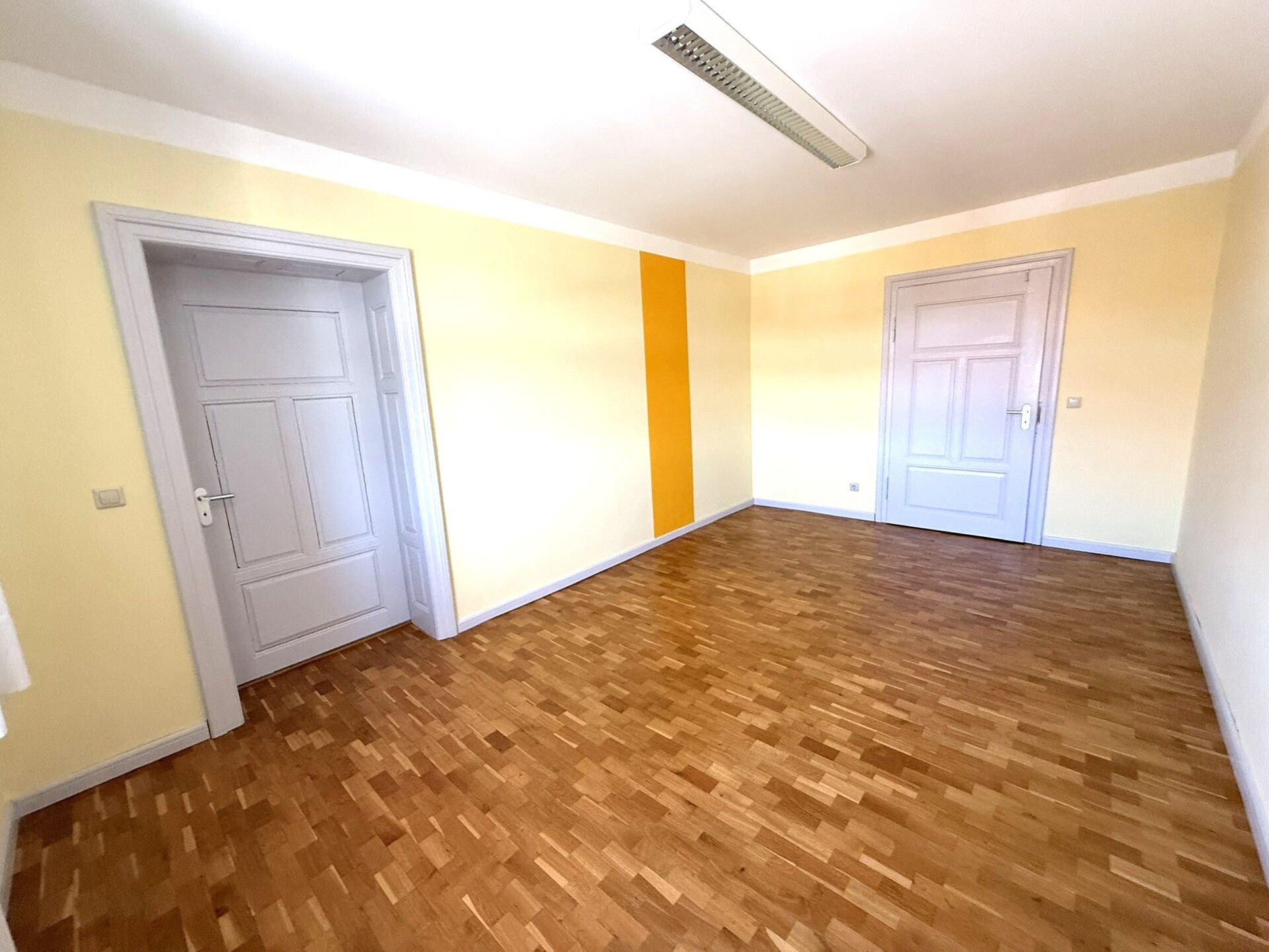 Wohnraum mit hellem Parkett, weißer Tür und gelber Akzentwand.