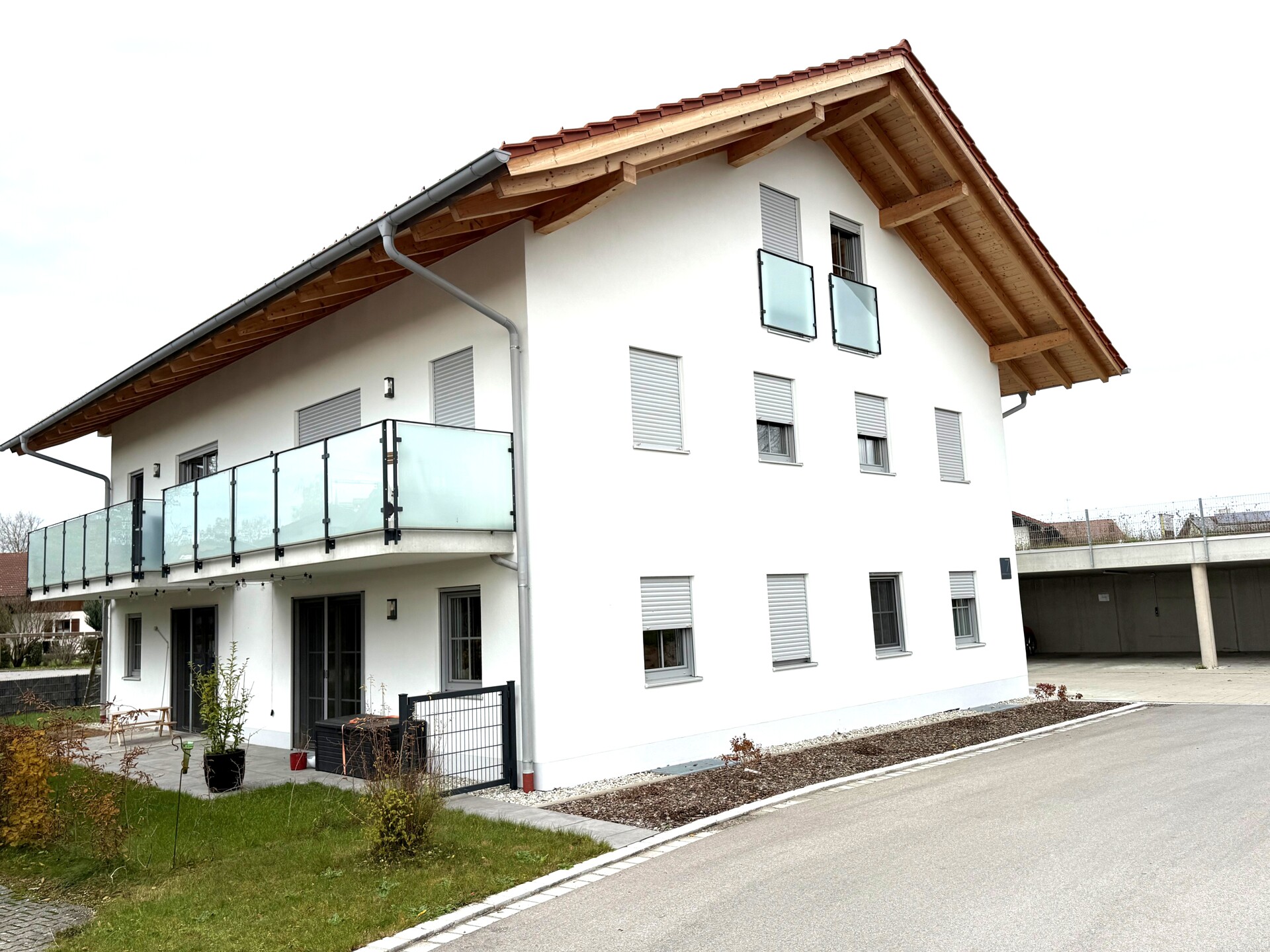 Außenansicht eines modernen, weiß verputzten Mehrfamilienhauses mit Holzgiebel, Balkon mit Glasbrüstung und großzügiger Einfahrt.