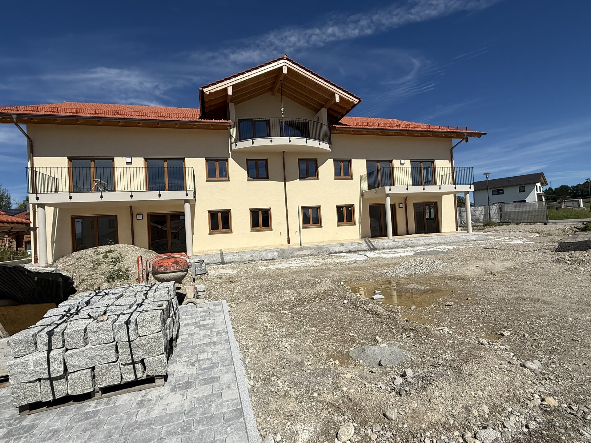 Neubau eines mehrstöckigen Wohnhauses mit hellgelber Fassade, rotem Satteldach und breitem Balkon, aufgenommen von der Baustelle mit Kiesfläche und Baumaterial im Vordergrund unter blauem Himmel.