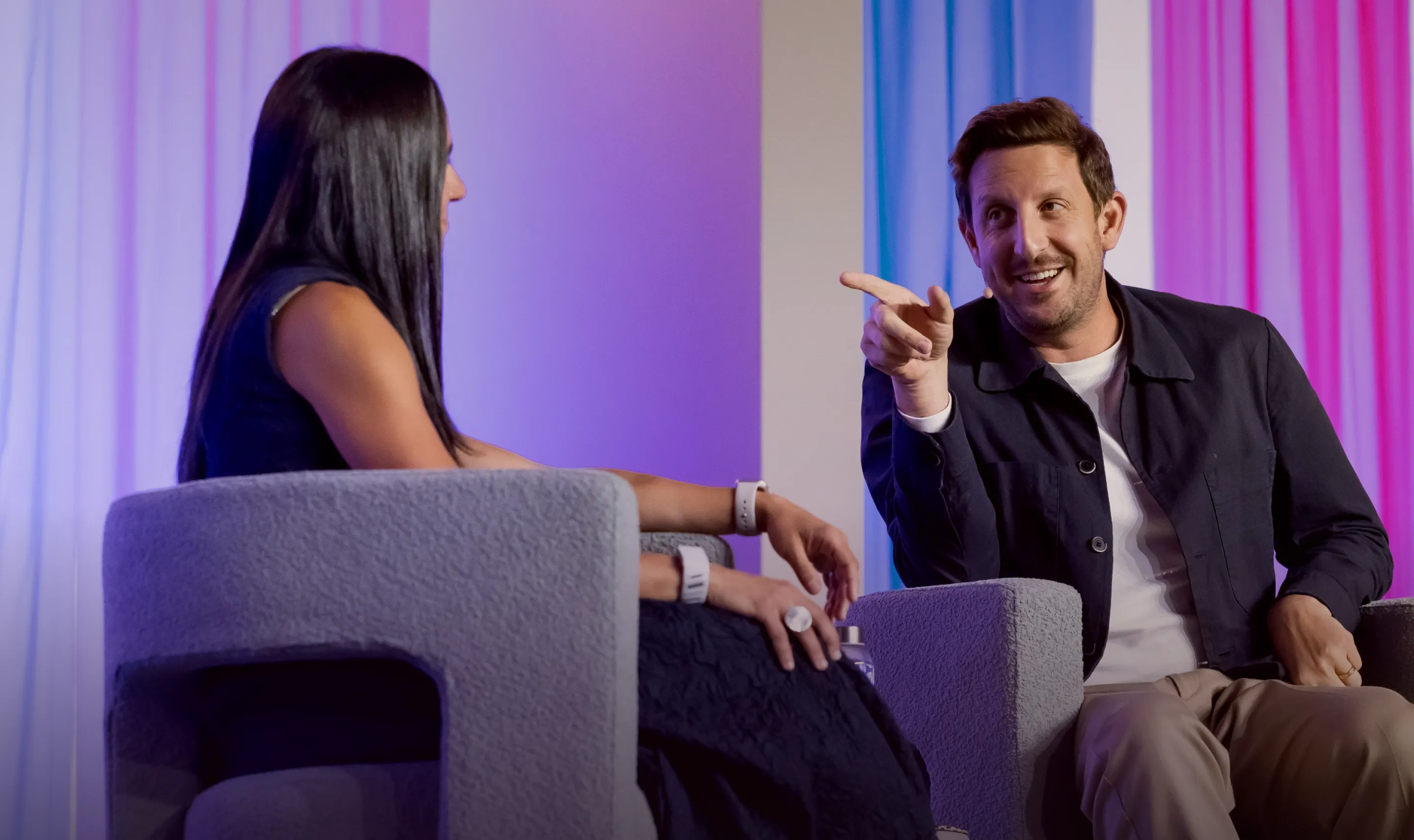 Man smiling and pointing while talking to a woman sitting in gray armchairs against a backdrop of pink and blue curtains.