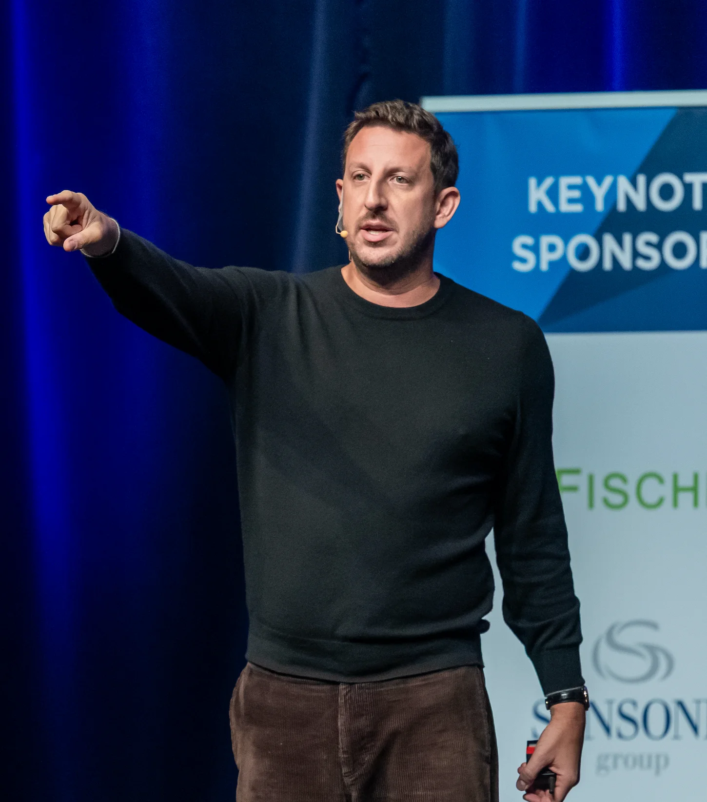 Man in black sweater speaking on stage with headset microphone, pointing to the left.